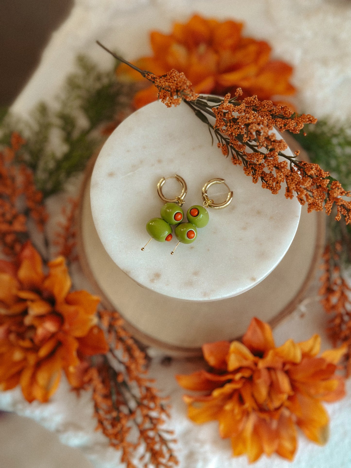 Handmade polymer clay martini olive earrings with green olives and red pimento detail on gold huggie hoops, styled with autumn flowers
