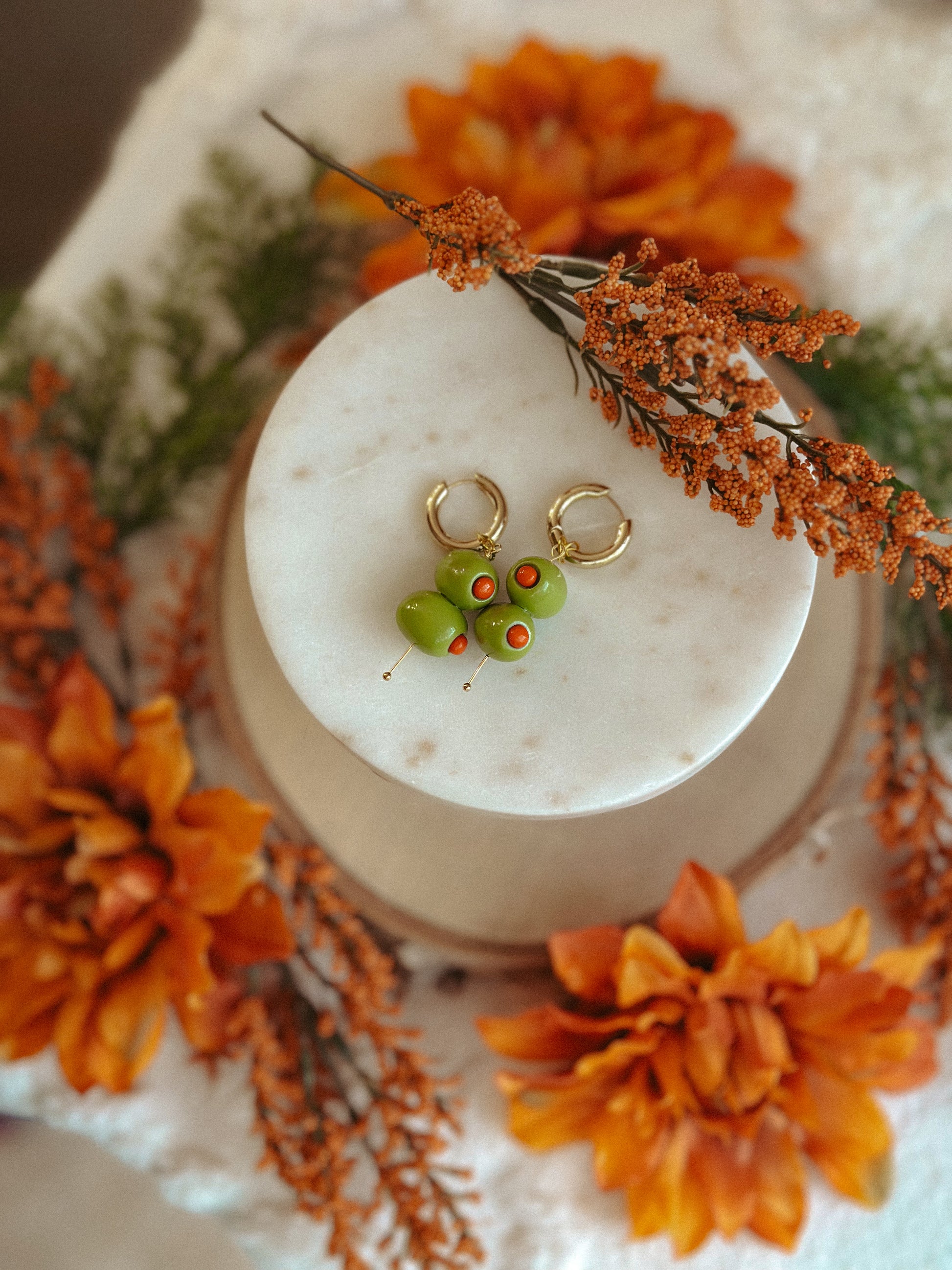 Handmade polymer clay martini olive earrings with green olives and red pimento detail on gold huggie hoops, styled with autumn flowers