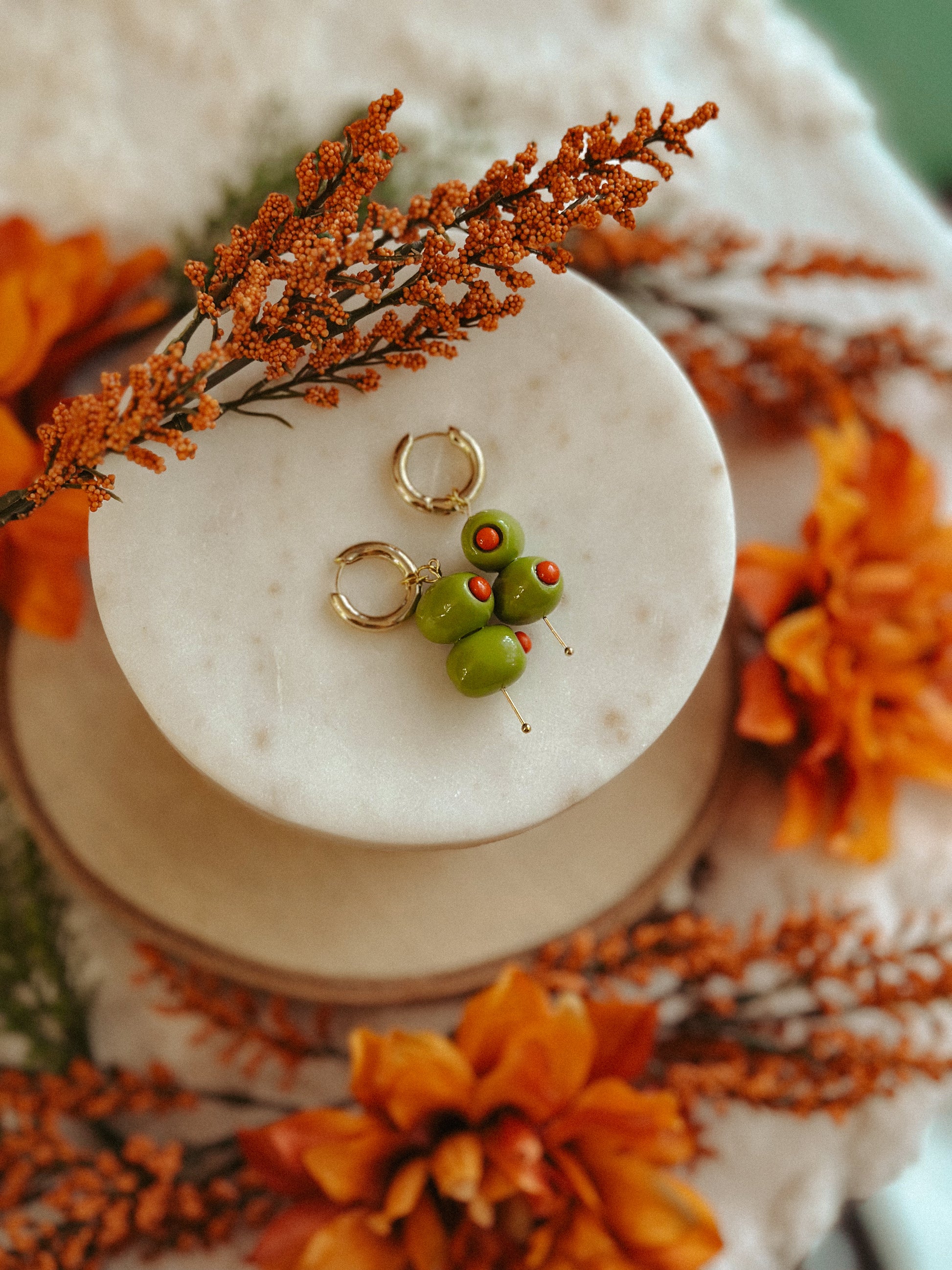 Handmade polymer clay martini olive earrings with green olives and red pimento detail on gold huggie hoops, styled with autumn flowers