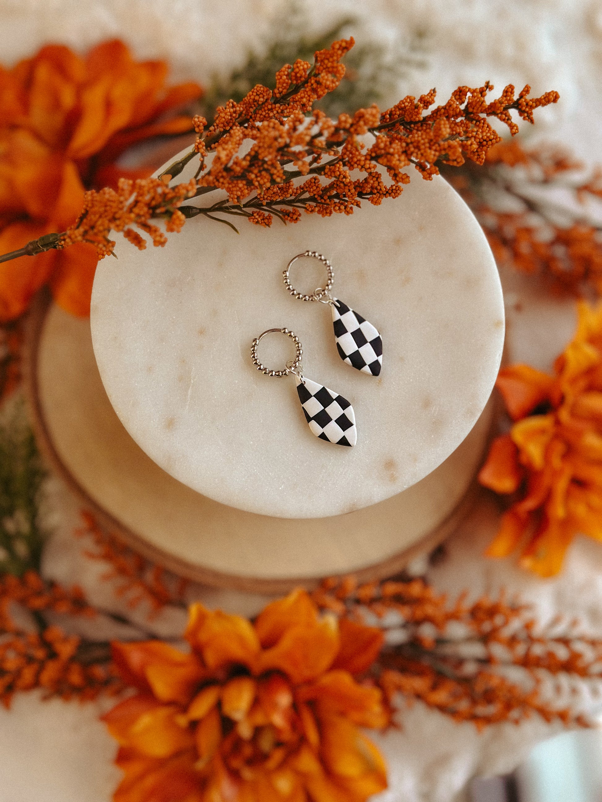 Black and white checkerboard teardrop earrings on beaded hoops, displayed on a round marble slab surrounded by autumn florals – handmade polymer clay jewelry by Olive & Arrow