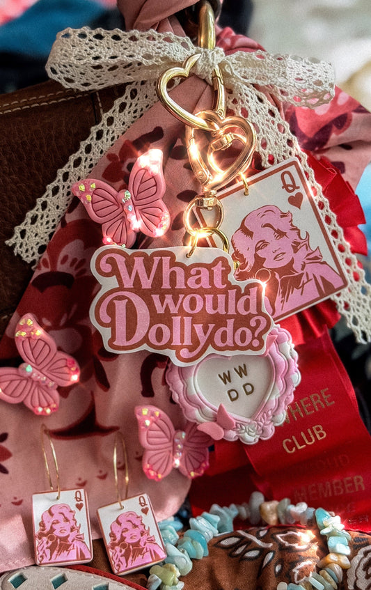 What Would Dolly Do pink script bag charm with gold heart clasp styled with Dolly-inspired butterfly and Queen card accessories
