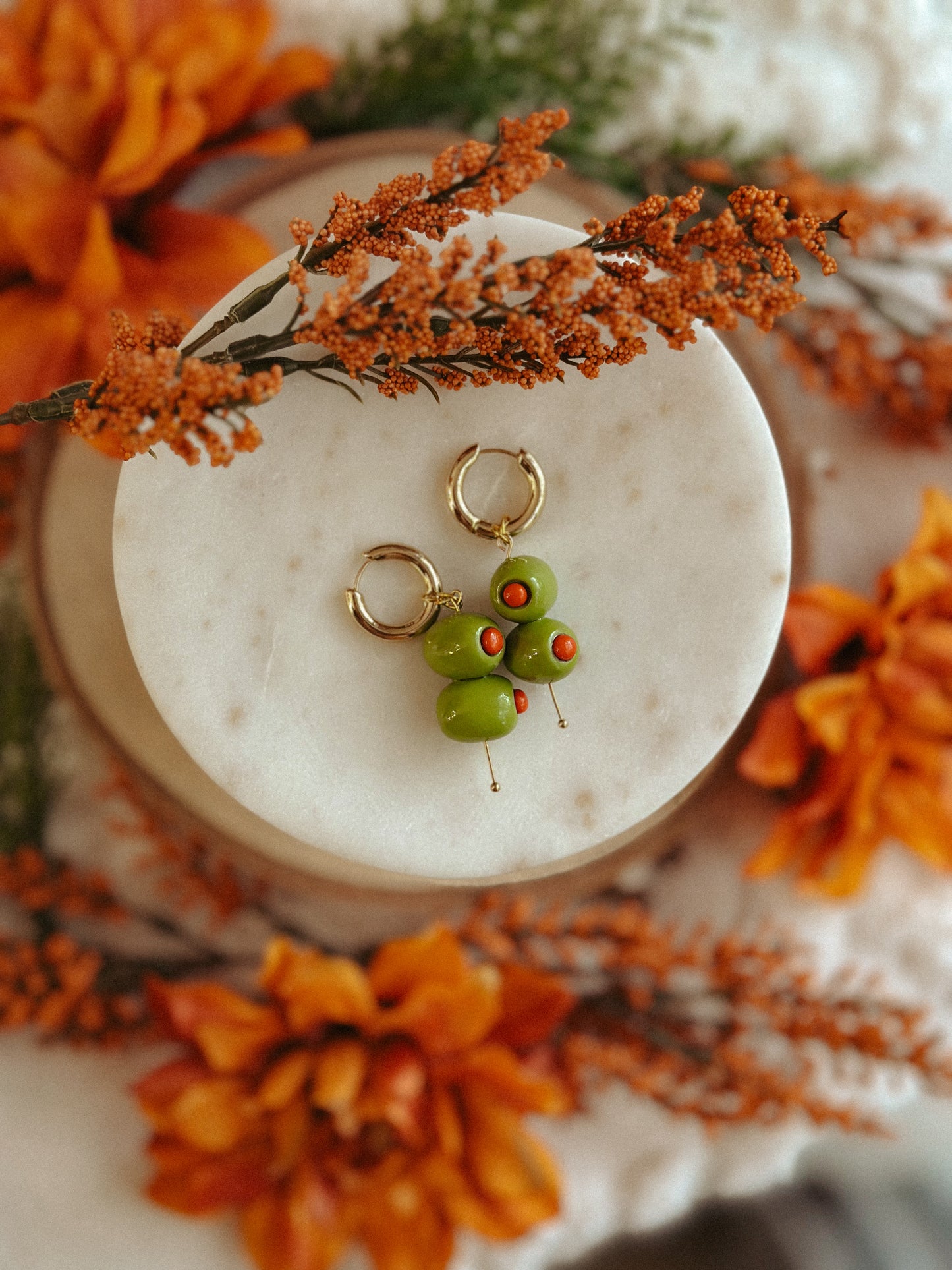 Handmade polymer clay martini olive earrings with green olives and red pimento detail on gold huggie hoops, styled with autumn flowers