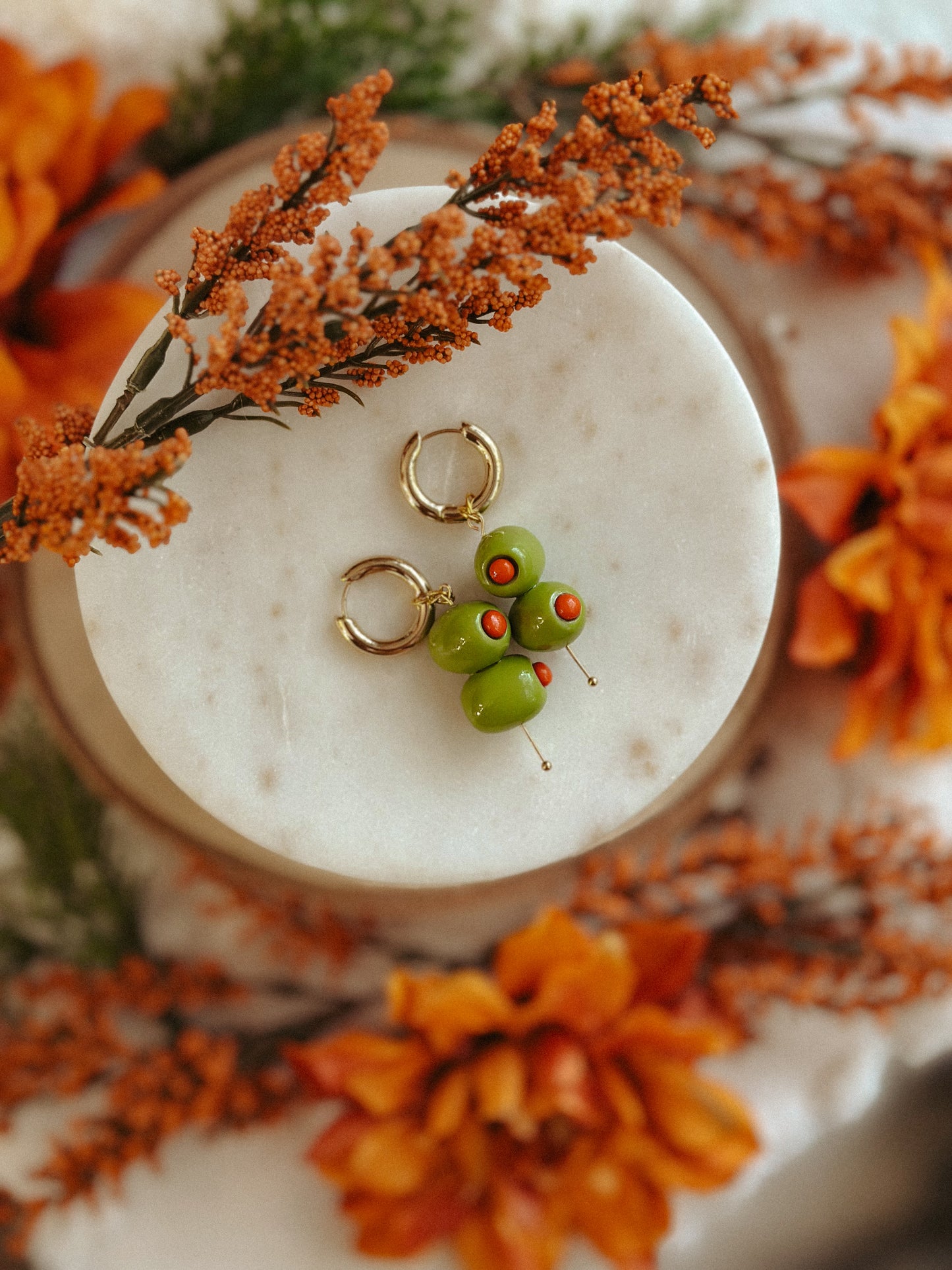 Handmade polymer clay martini olive earrings with green olives and red pimento detail on gold huggie hoops, styled with autumn flowers