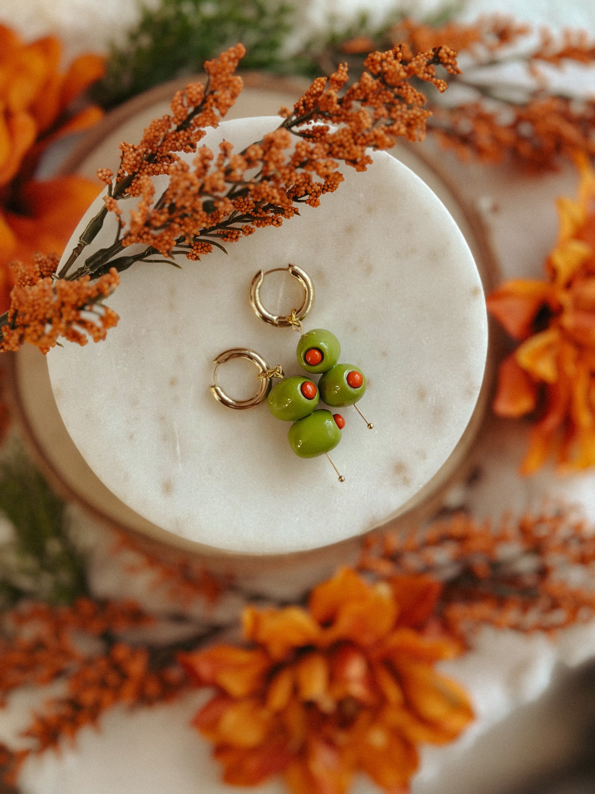 Handmade polymer clay martini olive earrings with green olives and red pimento detail on gold huggie hoops, styled with autumn flowers