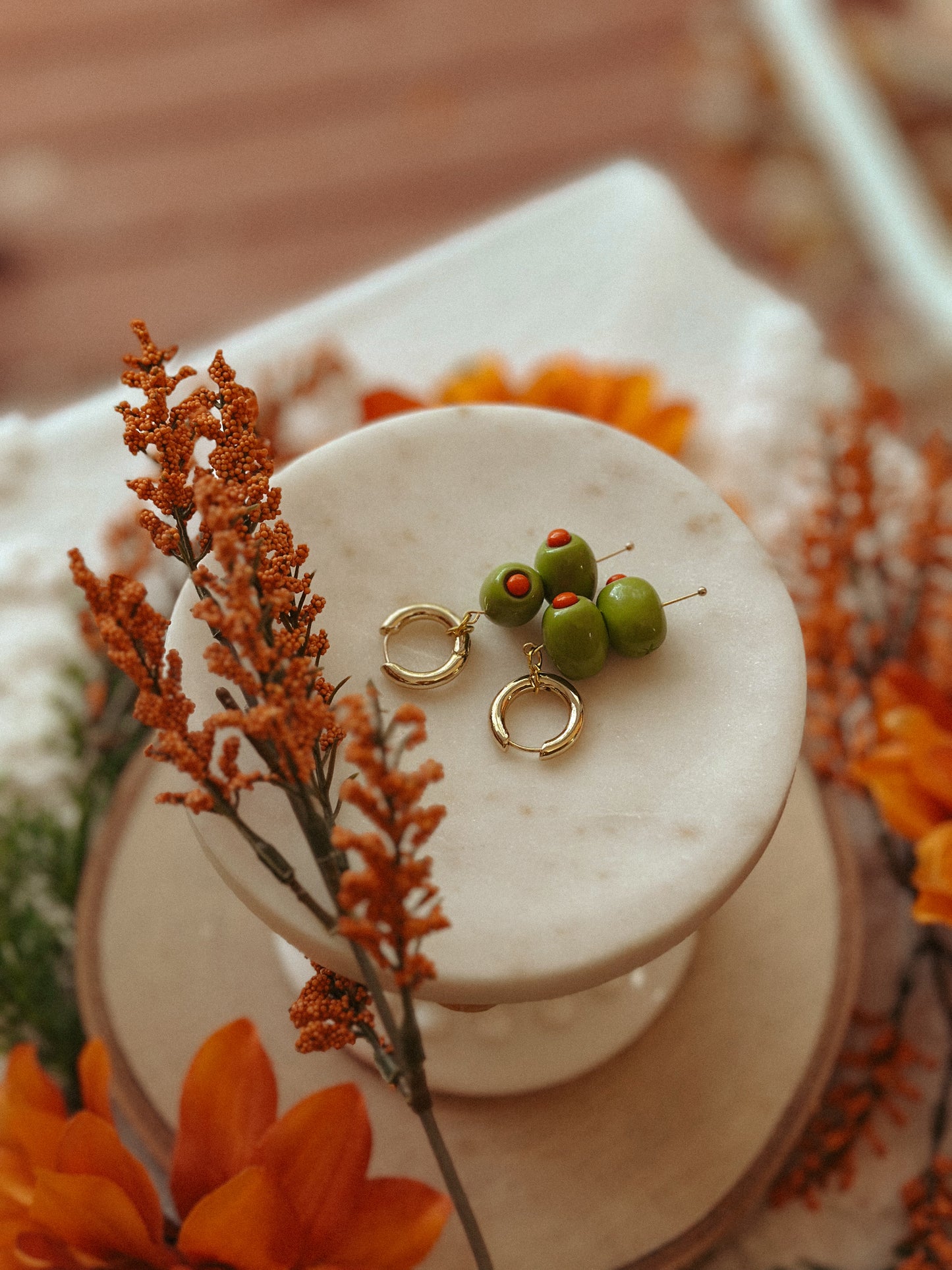 Handmade polymer clay martini olive earrings with green olives and red pimento detail on gold huggie hoops, styled with autumn flowers