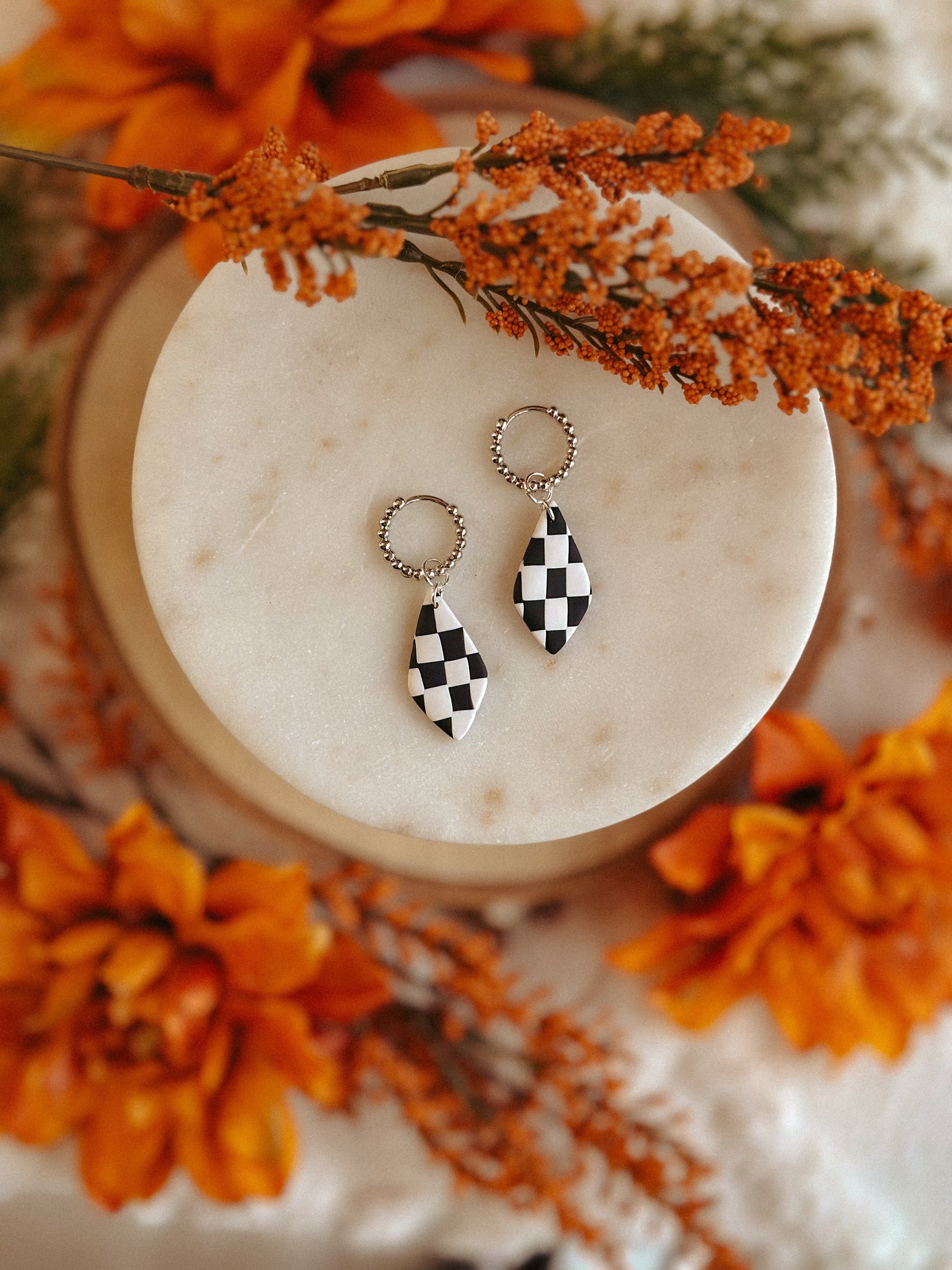 Black and white checkerboard teardrop earrings on beaded hoops, displayed on a round marble slab surrounded by autumn florals – handmade polymer clay jewelry by Olive & Arrow